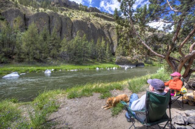 Two people sit riverside in camping chairs