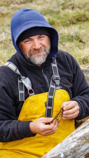 A man wearing yellow rubber overalls sitting in the grass holding a fox mandible