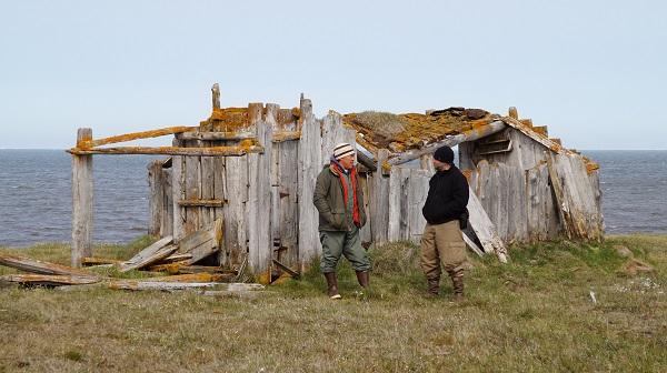Two men stand in front of a sod home built of drift wood and whale bones with the Arctic ocean in the background