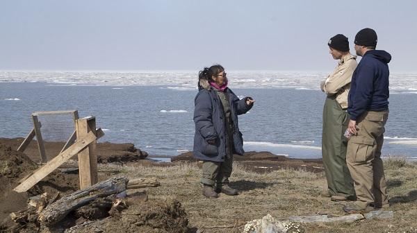 A woman speaking to two men, All are standing near an excavation site with the Arctic Ocean in the background