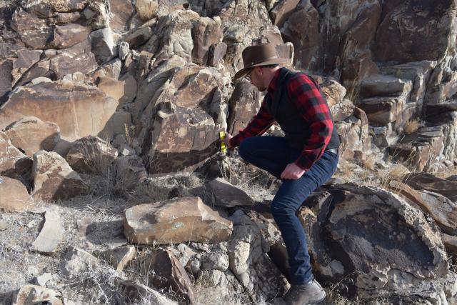 A person wearing a hat and a plaid shirt measures something with a small ruleron a rock outcropping.