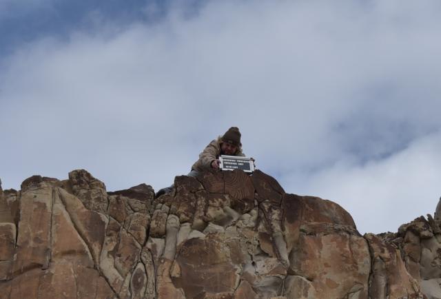 An individual wearing a hat is on top of a large rock outcropping holding a sign that reads Hogup with numbers to help document the site. There is a rock art site visible below the sign. 