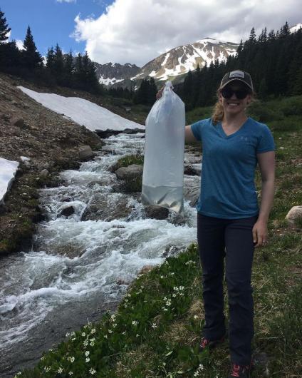 kelly standing near a creek with trees in the background holding a bag of fish