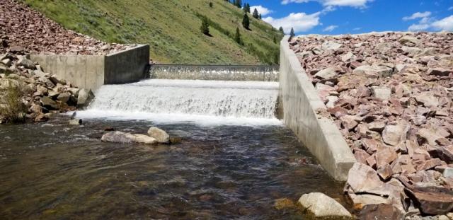 concrete fish barrier in creek. water flowing. rocks on either side. green hill in the background.