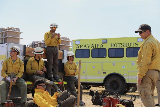 a group of firefighters eat lunch. A truck in the background reads Aravaipa Hotshots.