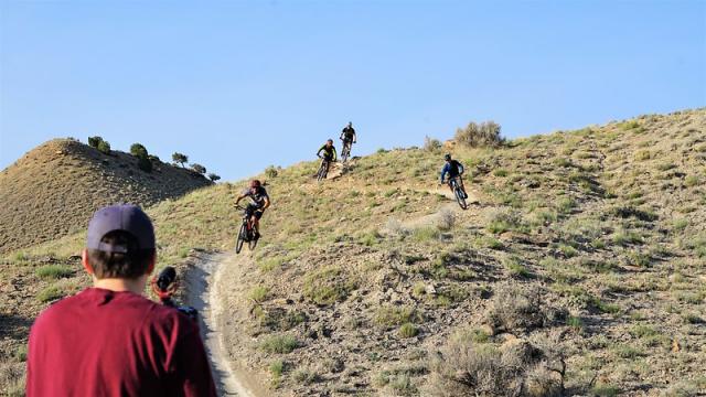 Man in red shirt films mountain bikers as they ride down a slope
