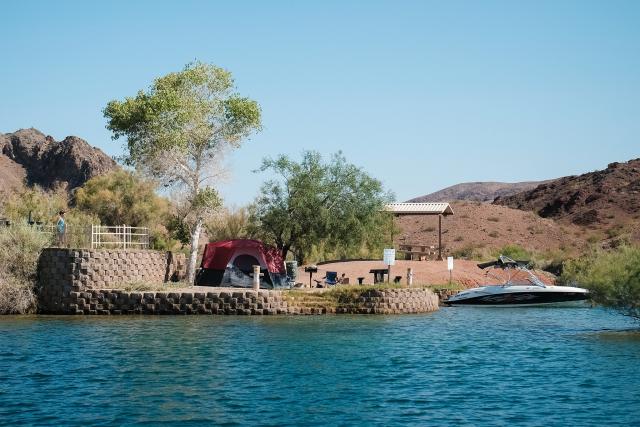 Campsite on a shoreline with a tent, a person standing above it on a constructed viewing area and a boat tethered nearby.