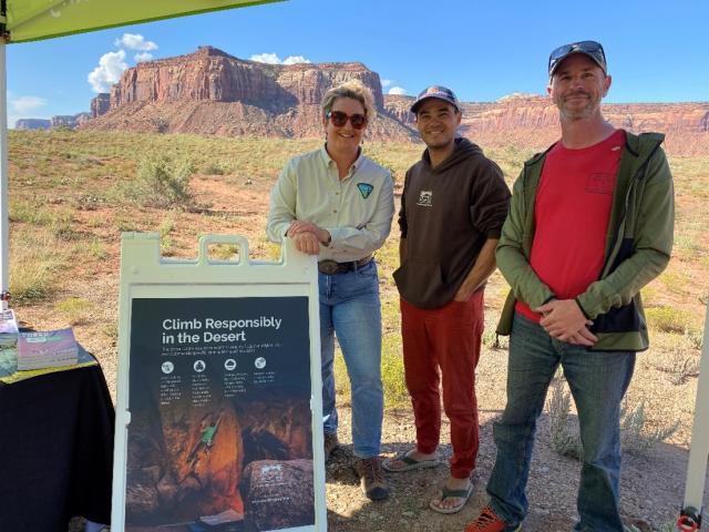 Three individuals under a canopy standing in behind a sign that says climb responsibly in the Desert. There are remarkable landscape of cliffs, clouds, and sky in the background.
