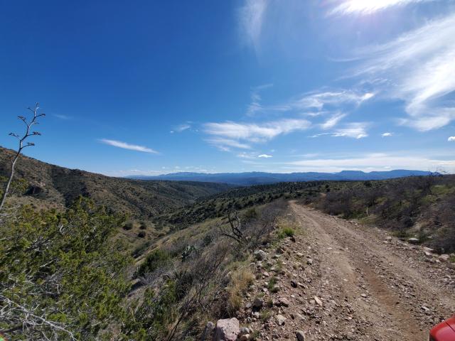 looking down a dirt road with beautiful mountains in the distance and a canyon on the left side. There is a very blue sky overhead with quite a few white wispy clouds
