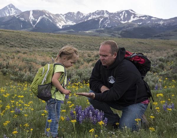 A man showing some seed to a girl surrounded by yellow and purple flowers. 