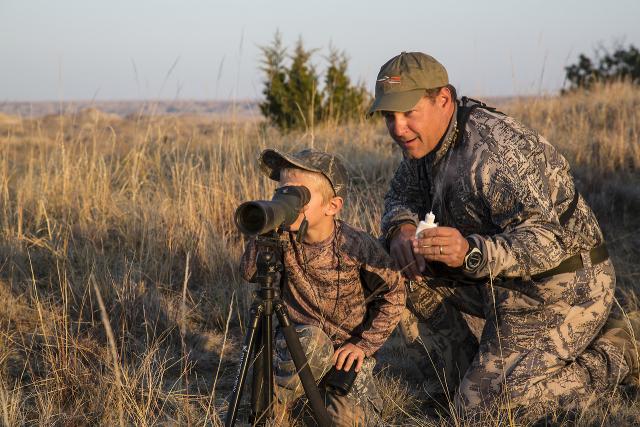 Father and son wearing camo gear, using a spotting scope