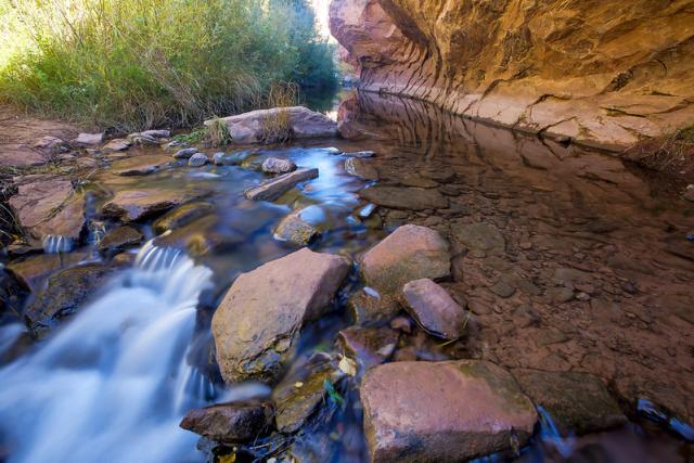 Water in a stream along Grandstaff Trail with red rocks showing through the clear water. A large rock cliff is on one side of the stream, the other side has green riparian plants continuing down the canyon.  Photo by Bob Wick