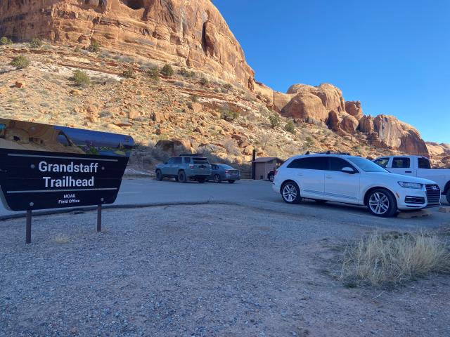 A shaded parking area with a sunny hill in the background. Cars sit in a parking lot at the trailhead.