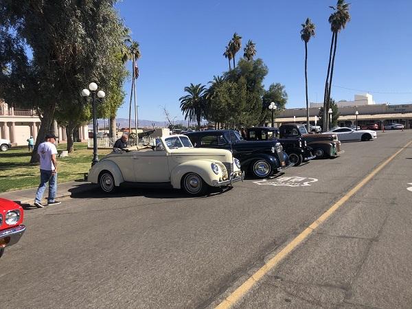 A white car, two black cars, and a brown car (all classic cars) parked along Route 66 and surrounded by trees.