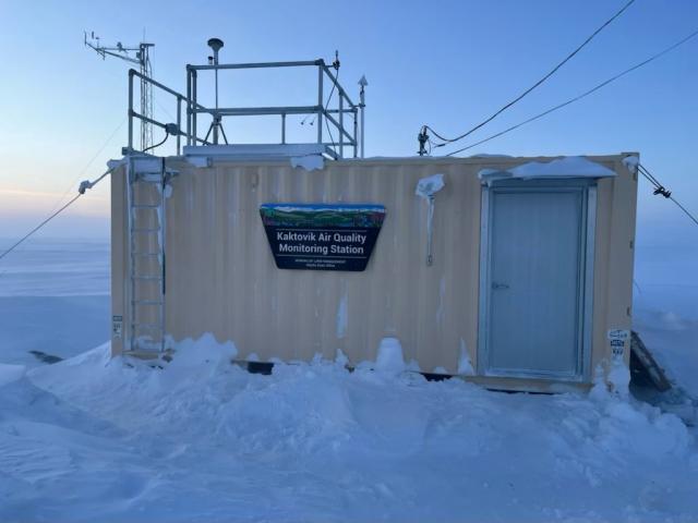 A shipping container with equipment sticking out of it. It has a trapezoid shaped sign that says "Bureau of Land Management Kaktovik Air Quality Monitoring Site."