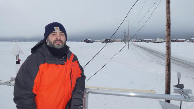 A man in an orange parka stands on top of a shipping container that houses air quality monitoring equipment. The Alaska Native Village of Kaktovik is visible in the background.