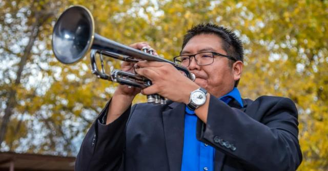 Delbert Anderson standing and playing the trumpet outdoors with trees behind him.