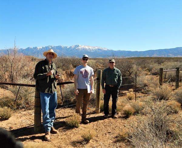 Three people standing in front of a fence with mountains in the background. 