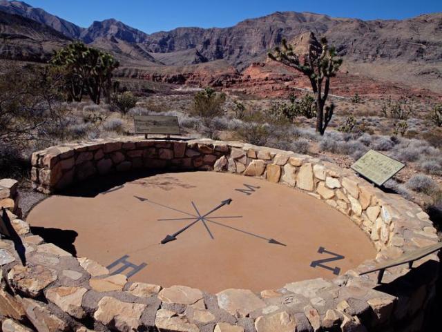 A very large compass surrounded by a stone wall with interpretive displays. Joshua trees and mountains are behind it.