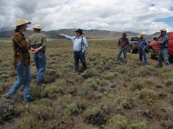 Ranchers standing in a circle outside with a gray and red truck in the background.