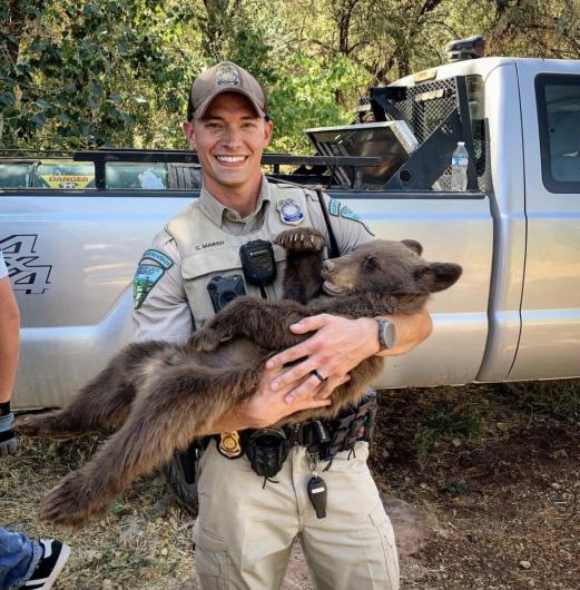 A BLM uniformed ranger standing in front of a truck with a bear cub in his arms. 