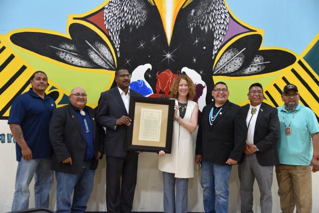 Bears Ears National Monument Inter-Governmental Cooperative Agreement signatories standing in front of a mural at the White Mesa Community Center smiling on June 18, 2022. They are holding the Presidential Proclamation restoring Bears Ears National Monument that was signed by President Biden on October 8, 2021.