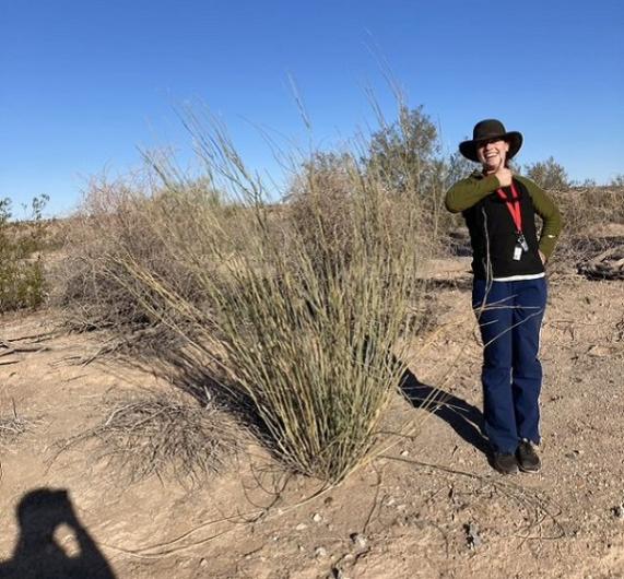 Botanist Maya Canapary stands next to a bush and gives the thumbs up