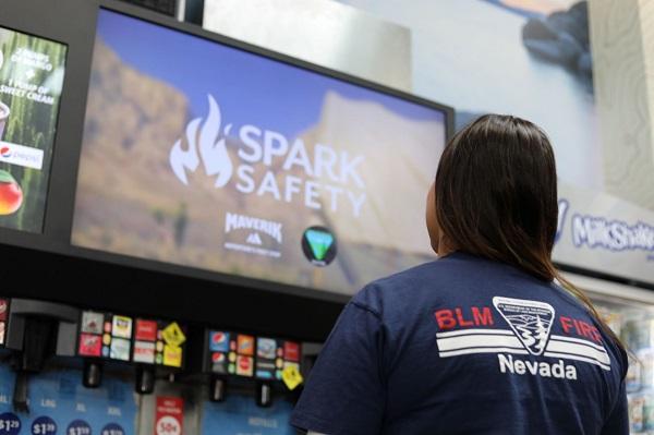 A woman looks up at the Spark Safety fire prevention messaging on monitors above fountain drinks. She is wearing a BLM Fire t-shirt. 