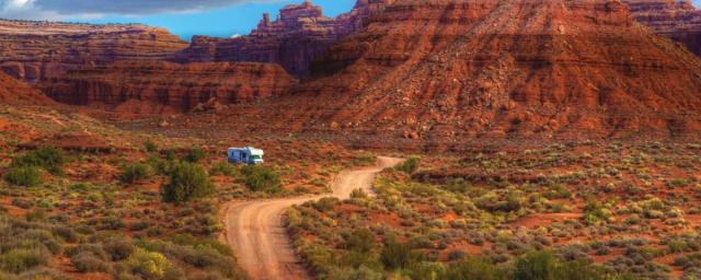 An RV driving on a dirt road through the Monticello River - San Juan area and red rock formations in the background. 