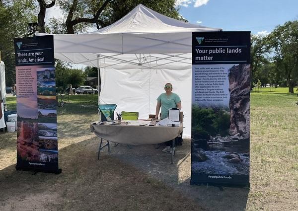 Marquessa Brown, BLM Human Resources Specialist, hosts the BLM information booth. She is standing under a white tent next to BLM info panels.