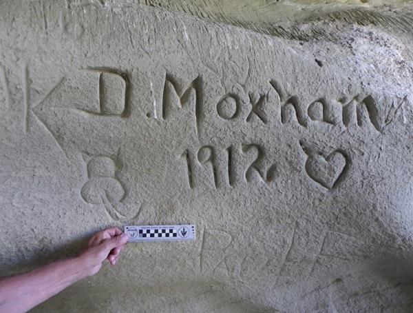 A volunteer holds up a measuring stick to a historic inscription that is on a rock face. 