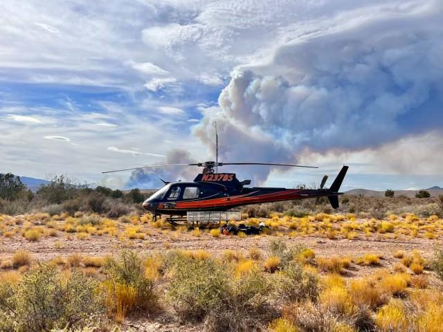 Helicopter sits stationary in a desert lands scape with short vegetation in the area (helispot). A large plume of smoke is visible behind the helicopter in the distance. 