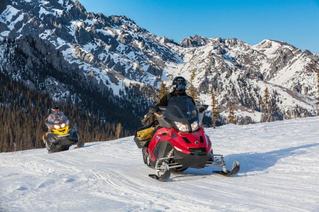 A red and yellow snowmobile riding upslope in front of a snow covered, rocky slope on a blue-sky day.