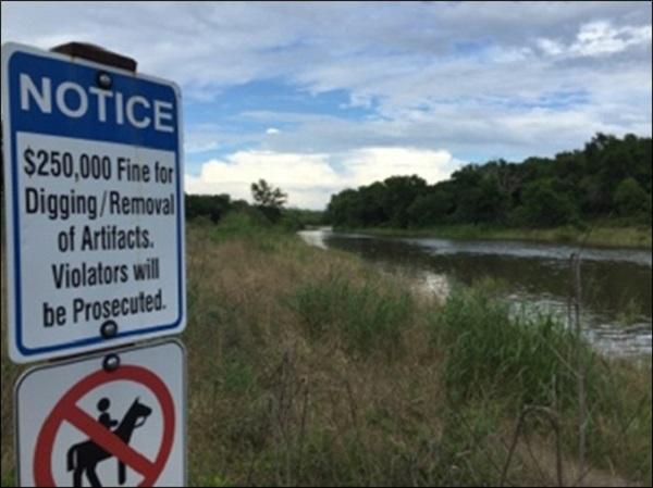 A warning sign that says: "Notice: $250,000 fine for digging/removal of artifacts. Violators will be prosecuted. Below that sign is another sign of a person on a horse crossed out. Next to the signs is a stream. 