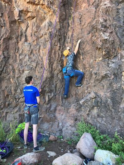 A student is climbing the rock face while a staff member below is holding a purple rope and watching the student. 