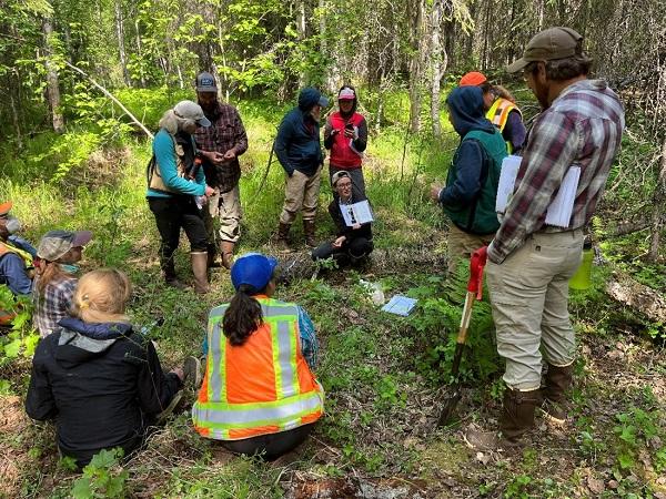 Ruth Whittington of the Colorado Natural Heritage Program helps teach Riparian and Wetland AIM crews about soil structure. Participants are standing around in a forest. 