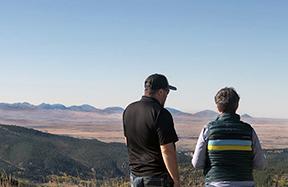 A man (left) and woman (right) conversing (viewed from the back), facing toward a far-reaching landscape of hills, a valley prairie and distant mountain range.
