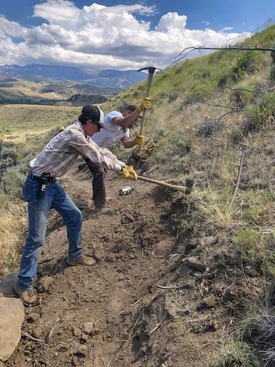 Two MCC crew members using tools to pound the side of the dirt trail. 