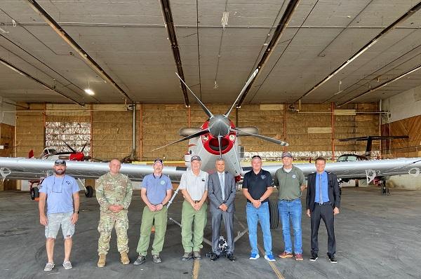 Eight members of the group stand in front of a plane.