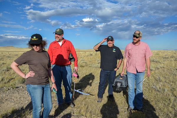 A group of four people stand in a field with Cody Field Manager Cade Powell holding a ferret in a cage. 