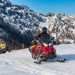 A person straddles a red snowmachine and rides across snowpack. Mountains and blue sky in background.