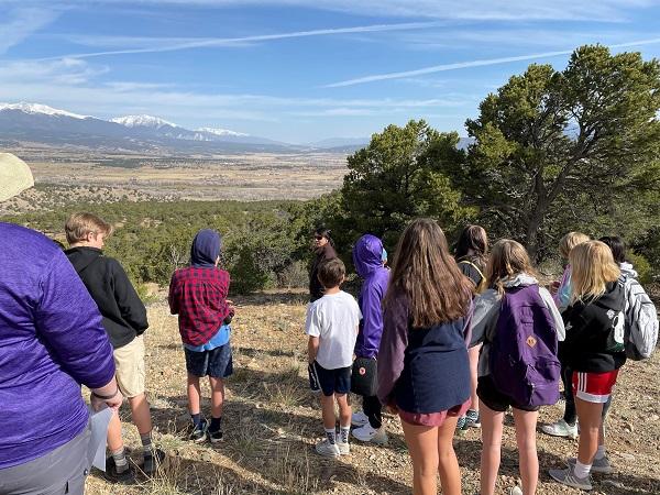 A group of middle school students is standing with a BLM Natural Resource Specialist and looking out over the Colorado landscape. Mountains are in the distance. 