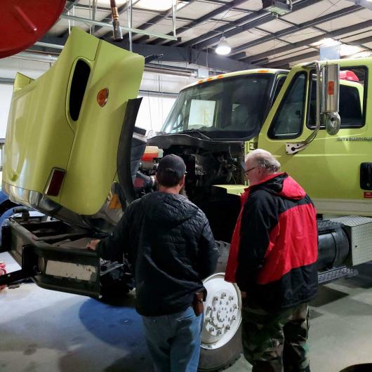 Two individuals stand in front of a truck with the hood up, looking inside. The truck was transferred through the rural fire readiness program.