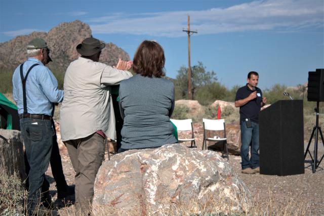 man standing outside behind a podium with two people standing and one sitting on a rock listening to him talk