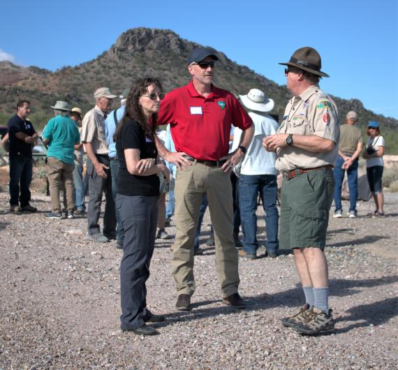 Three people standing and talking with a crowd of people behind them and a mountain in the background.