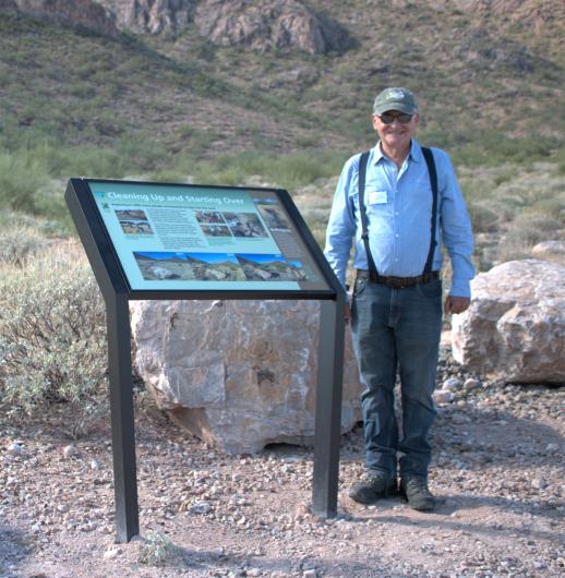 Person wearing suspenders and a ball cap standing next to an interpretive panel