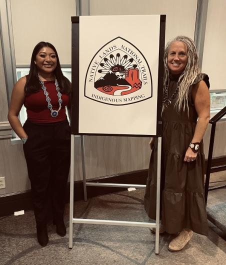 Kiana Etsate-Gashytewa (left) with BLM National Scenic and Historic Trails (NSHT) Program Lead, Carin Farley (right). They are standing next to a poster of the Indigenous Mapping and Research Project (IMRP) logo.