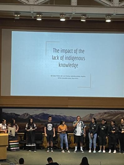 Group of people standing on a stage with presentation on a screen overhead that says the impact of lack of indigenous knowledge.