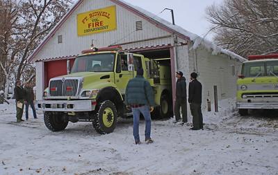 A green fire engine in a white garage with snow on the ground and roof. People are standing outside the garage. 