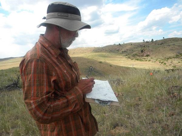 Ben holding a pen and writing on a clipboard in the middle of grassy hills. 